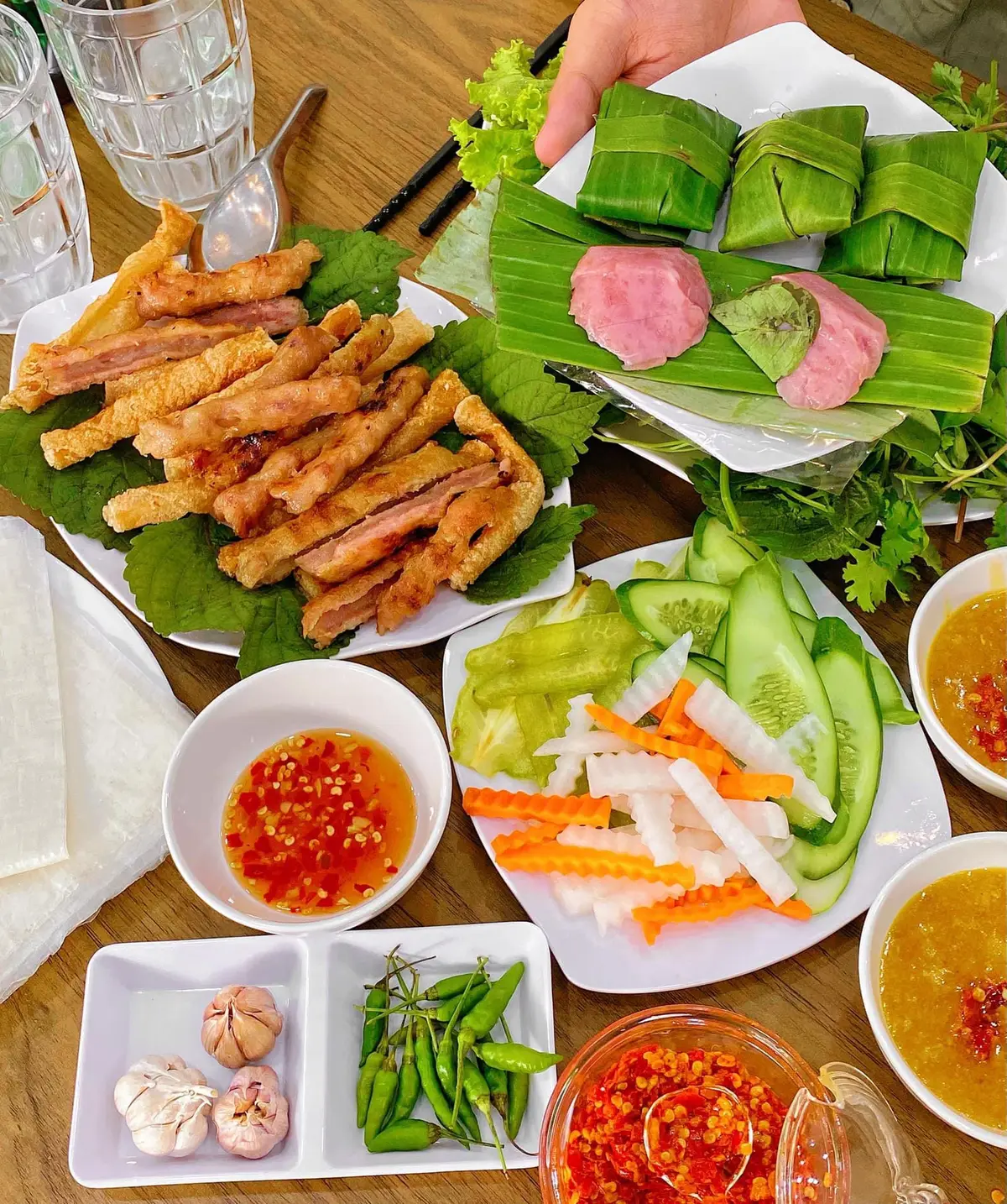 Table with grilled pork skewers, fried rolls, sliced vegetables and two dipping sauces